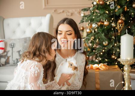 Eine Mutter und ihre Tochter genießen einen besonderen Moment zusammen, während sie mit einem Kaninchen neben einem wunderschön geschmückten Weihnachtsbaum interagieren. Sunligh Stockfoto