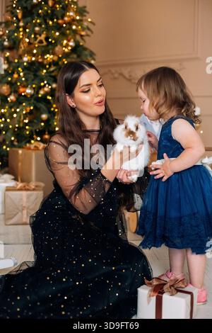Eine fröhliche Mutter hält einen kleinen Welpen, während ihre Tochter sich hinlehnt, um ihn zu küssen. Sie sind umgeben von wunderschön verpackten Geschenken und einem glitzernden Weihnachtsfest Stockfoto