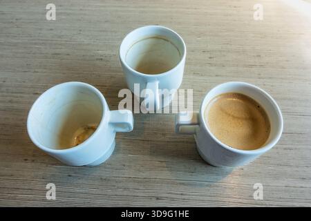 Drei weiße Keramikbecher auf einem Holztisch von oben, einer mit frischem Espresso gefüllt, zwei mit gebrauchtem Kaffeepause, was auf eine Kaffeepause oder ca. Stockfoto