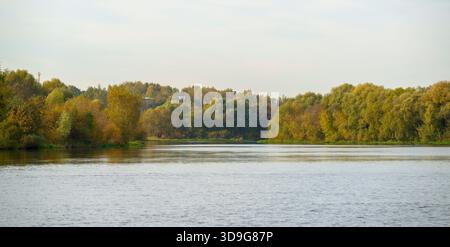 Weite Aussicht auf einen ruhigen Fluss, der sich zwischen herbstfarbenen Bäumen unter hellem Himmel erstreckt. Ruhige, saisonale Landschaft mit natürlichen Reflexen und sanftem Licht Stockfoto