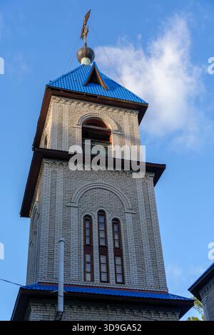 Vertikaler Blick auf einen kunstvoll verzierten orthodoxen Glockenturm mit blauem Metalldach und Kreuz vor einem hellen Himmel mit hellen Wolken bei natürlichem Tageslicht. Hohe qu Stockfoto