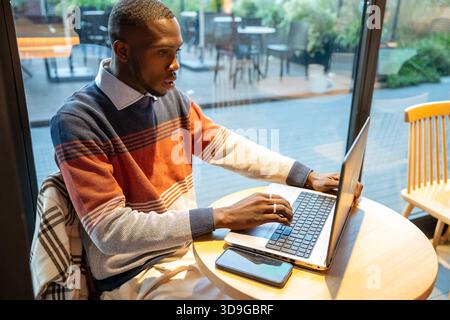 Ein junger Mann sitzt an einem runden Holztisch und tippt auf einem Laptop in einem warmen und einladenden Café. Er trägt einen farbenfrohen Pullover und konzentriert sich tief, umrahmt Stockfoto