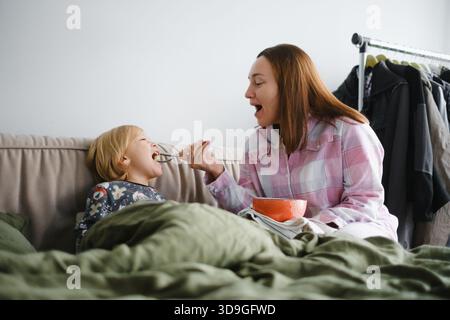Mutter und Sohn frühstücken zusammen oder essen im Schlafanzug im Bett, Familienurlaub beim Frühstück im gemütlichen Schlafzimmer mit Kissen Stockfoto