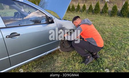 Ein Mann in einer orange-grauen Jacke, der das rechte Vorderrad eines geparkten silbernen Autos mit offener Motorhaube auf einer grasbewachsenen Straße überprüft. Stockfoto