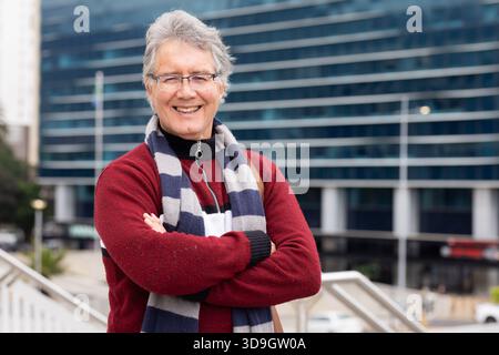 Ein älterer Mann, der auf dem Stadtrand steht, lächelnd mit Brille, gestreiftem Schal neben einem Metallgeländer Stockfoto