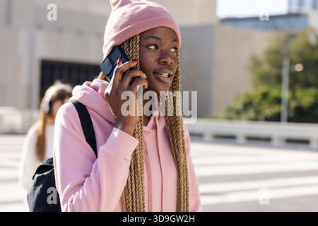 Afroamerikanerin im Kapuzenpullover, die am Straßenübergang der Stadt steht und am Telefon spricht und einen Rucksack trägt Stockfoto