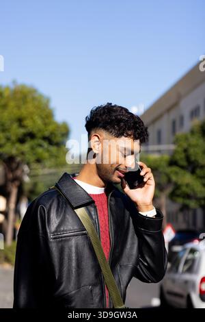 Indischer Mann in 20ern, der auf dem Smartphone auf der Stadtstraße spricht, in Lederjacke, Umhängetasche, Armbanduhr Stockfoto