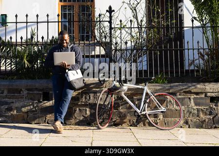 Afroamerikaner lehnt sich an eine Steinmauer auf dem Bürgersteig und schreibt auf Laptop mit dem Fahrrad, Kopierraum Stockfoto