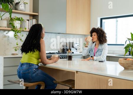 Verschiedene Freundinnen unterhalten sich, während sie Kaffee aus Tassen in der Nähe der französischen Presse auf der Kücheninsel schlürfen Stockfoto
