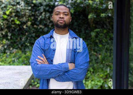 Afroamerikaner, der mit überkreuzten Armen steht und Denim-Hemd auf Betonbank inmitten der Efeuwand trägt Stockfoto
