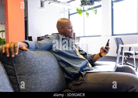 Afroamerikaner sitzt auf grauem Sofa in der Bürolounge und hält Smartphone und Laptop offen Stockfoto