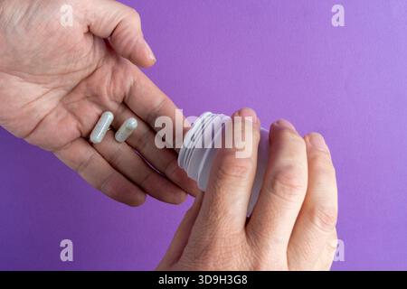 Nahaufnahme der Hände, die weiße Kapseln aus einer Flasche auf die Palme gießen, die Gesundheit oder Medikamente auf einem lila Hintergrund symbolisieren. Stockfoto