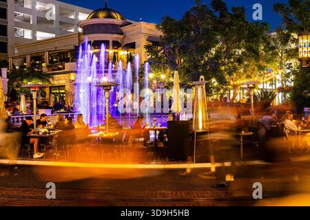 Shopper und Gastronomen im Grove at Farmers Market in Los Angeles, Kalifornien, machen einen Spaziergang durch ein Einkaufszentrum im Freien, vorbei am Brunnen bei Nacht, lange Zeit ausgesetzt Stockfoto