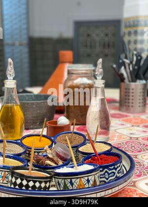 Farbenfrohe marokkanische Gewürze in der traditionellen Küche von Marrakesch, in gemusterten Schalen auf einem Tisch mit Tagine, Ölflaschen und Utensilien Stockfoto