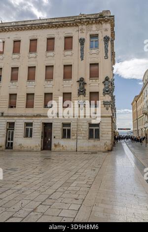 SPLIT, KROATIEN, 10.25.2025: Ein verwittertes, kunstvolles Gebäude säumt eine sonnenbeschienene Uferstraße. Skulpturale Reliefs zieren die Ecke, während geschlossene Fensterläden Hi Stockfoto