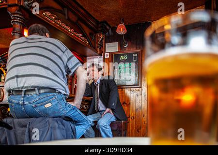 Zwei Männer sitzen in einem Pub in London. Ein Mann trägt einen Anzug, der andere ein gestreiftes Hemd. Ein Pint Bier steht auf dem Tisch. Stockfoto