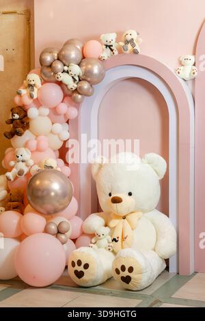 Das Foto zeigt eine dekorative Einrichtung mit einem großen Plüschteddybär, Bogenelementen und einer Ballongirlande in sanften Rosa- und Goldtönen. Stockfoto