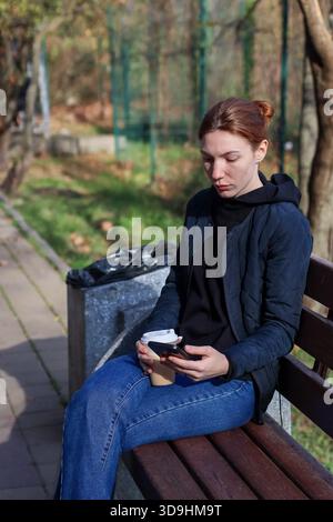 Junge Erwachsene Frau surft mit Kaffee in der Hand am Telefon, während sie sich auf einer hölzernen Parkbank im Freien entspannt Stockfoto