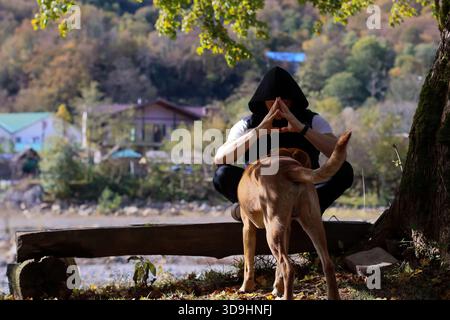 Junger Mann in schwarzem Hoodie sitzt im Kreuz auf Holzbank am Fluss, Hände im Gebet, brauner Hund neben ihm, Herbstlaub und Berge im Hinterland Stockfoto