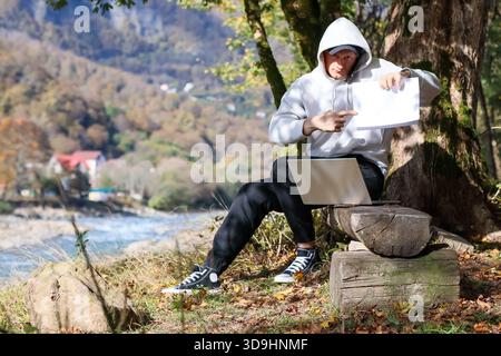 Ein lächelnder Mann im Kapuzenpullover zeigt Papierbogen und Gesten, während er auf einem Baumstamm neben dem Baum sitzt, mit Laptop im Herbst Stockfoto