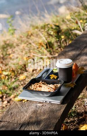Getreideschale in schwarzem Behälter mit Holzgabel, Kaffeetasse zum Mitnehmen, orangen Früchten, gelben Blättern auf silbernem Laptop auf Holzbank mit Blick auf W Stockfoto