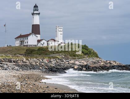 Montauk Point Lighthouse von der felsigen Atlantikküste von Long Island aus betrachtet, während die Wellen darunter brechen und Besucher das Gelände erkunden. Stockfoto
