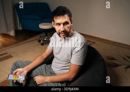 Ein bärtiger Mann, der Kopfhörer mit Knochenleitung trägt, entspannt sich zu Hause und konzentriert sich auf das Smartphone, während er Musik und Podcasts streamt, um privat und bequem zu sein Stockfoto
