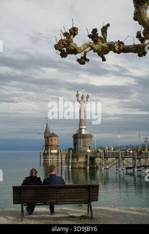 Zwei Personen sitzen auf einer Bank, entspannen sich und schauen sich die berühmte Imperia-Statue am Hafen von Konstanz am Bodensee an. Stockfoto