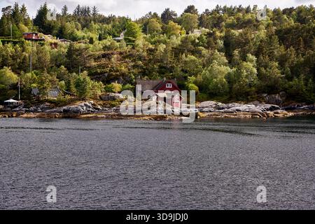 Ein winziger weißer Leuchtturm mit rotem Dach steht auf einer felsigen, bewaldeten Insel entlang eines schmalen norwegischen Fjords unter dramatischem bewölktem Himmel und weichem Nachmittag Stockfoto