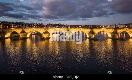 Die Aussicht zeigt die Karlsbrücke in Prag am Abend. Die Brücke ist beleuchtet und reflektiert über dem Wasser darunter. Boote sind auf dem Fluss, um die Szene zu verstärken. Stockfoto