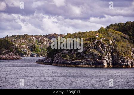 Ein winziger weißer Leuchtturm mit rotem Dach steht auf einer felsigen, bewaldeten Insel entlang eines schmalen norwegischen Fjords unter dramatischem bewölktem Himmel und weichem Nachmittag Stockfoto