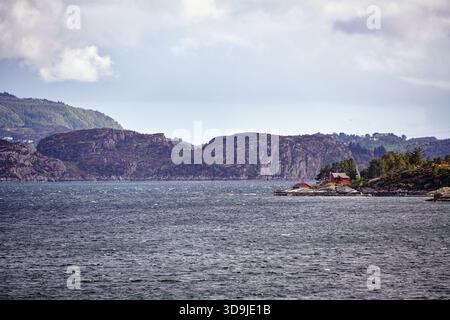 Ein winziger weißer Leuchtturm mit rotem Dach steht auf einer felsigen, bewaldeten Insel entlang eines schmalen norwegischen Fjords unter dramatischem bewölktem Himmel und weichem Nachmittag Stockfoto