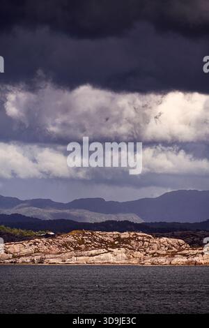 Ein winziger weißer Leuchtturm mit rotem Dach steht auf einer felsigen, bewaldeten Insel entlang eines schmalen norwegischen Fjords unter dramatischem bewölktem Himmel und weichem Nachmittag Stockfoto