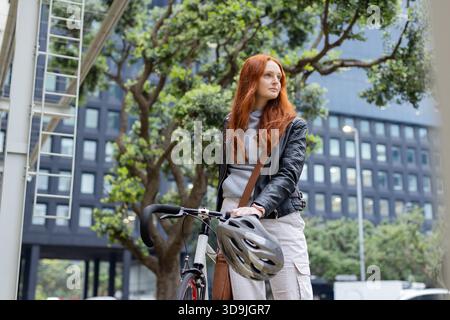 Rothaarige Radfahrerin, die am Fahrrad auf dem Bürgersteig steht und eine Lederjacke mit Helm und Tasche trägt Stockfoto