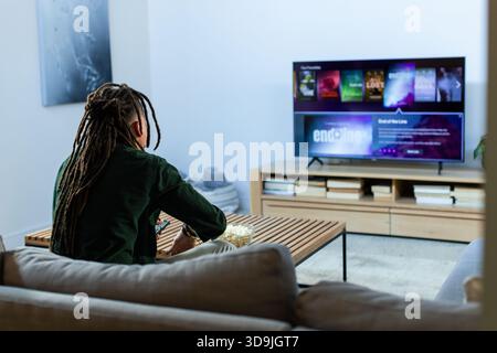 Person mit Dreadys auf dem Sofa sitzend auf dem eingebauten Fernseher mit Popcorn-Schüssel, Fernbedienung im Wohnzimmer Stockfoto