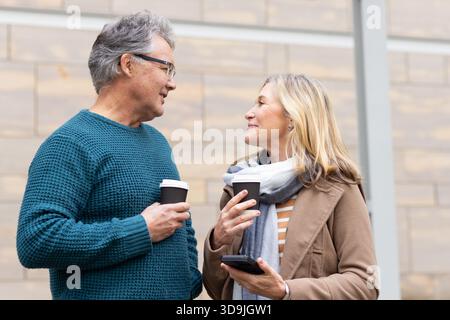 Seniorenpaar, das auf dem Bürgersteig vor der Steinfassade steht und Kaffeetassen hält und telefoniert Stockfoto