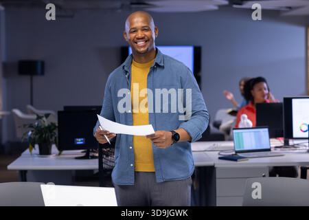 Afroamerikanischer männlicher Büroangestellter, der Dokumente hält, während er am Schreibtisch neben der Anlage steht Stockfoto
