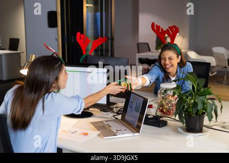 Verschiedene weibliche Kollegen lachen, während sie in einem modernen Büro am Schreibtisch ein Geschenk mit grüner Umhüllung austauschen Stockfoto