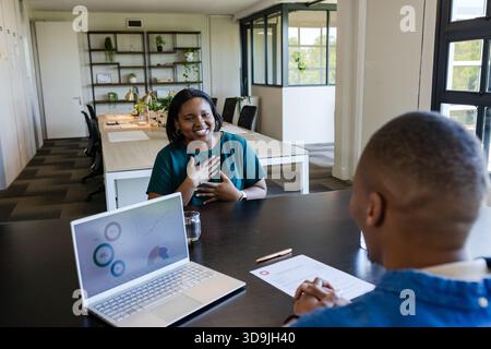 Afrikanische Kollegen sitzen im Büro, lächelnde Frau, die Hand an Brust hält, Laptop zeigt Karten Stockfoto