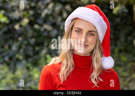 Frau, die im Garten mit grüner Hecke steht, mit rotem Rollkragenpullover und Weihnachtsmann-Hut Stockfoto
