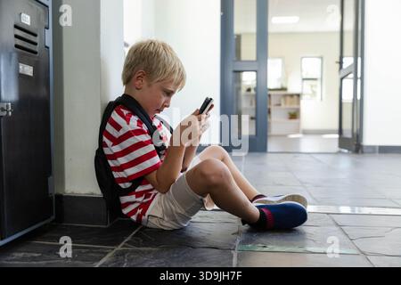 Junge im Schulalter, der in gestreiftem Hemd und Rucksack im Schulflur sitzt, mit Smartphone, Kopierraum Stockfoto