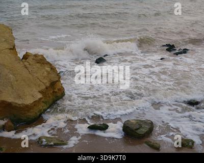 Nahaufnahme von mächtigen Meereswellen, die auf einen Sandstrand mit großen gelben und dunklen Felsen stürzen. Weißer Schaum, aufwirbelndes Wasser, Bewegung, natürliches abstraktes, roh Stockfoto
