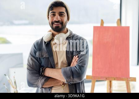 Indischer Mann mit Kopfhörern, stehend an der Staffelei im Kunststudio mit roter Leinwand, Pinsel Stockfoto