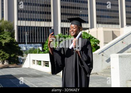Afroamerikanerin mit Mütze, die auf dem Campus posiert und Selfie mit Diplom und Smartphone macht Stockfoto