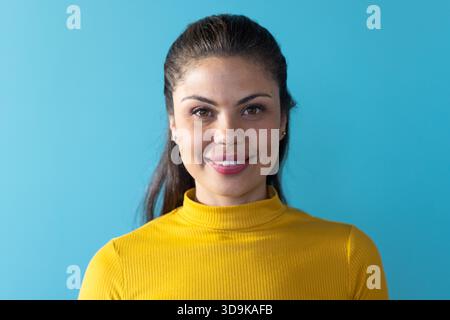 Frau in Senfpullover mit Ohrstecker, die vor blauem Hintergrund steht und im Studio lächelt Stockfoto