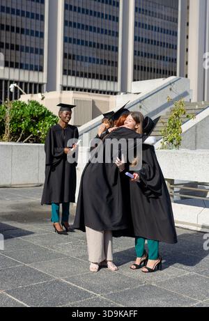 Verschiedene weibliche Absolventen in Kleidern und Mützen, die sich an der plaza umarmen, mit Pflanzgefäßen, die ein Rolldiploma halten Stockfoto