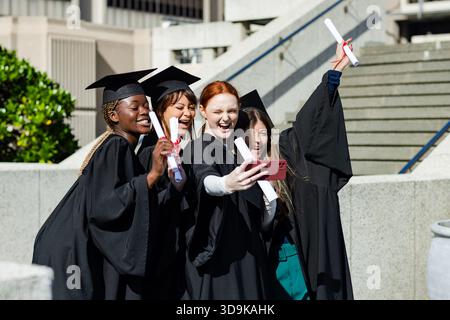 Verschiedene weibliche Absolventen posieren auf dem Campus in Caps mit Diplomen und machen Selfie mit Smartphone Stockfoto