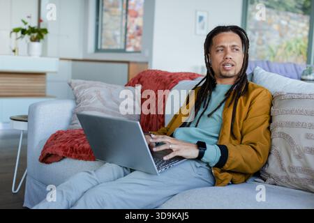 Afroamerikanischer Mann mit Senfjacke, der zu Hause auf dem Sofa sitzt und am Laptop arbeitet, Kopierraum Stockfoto