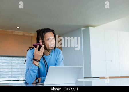 Afroamerikaner lehnt sich auf die weiße Kücheninsel-Arbeitsplatte, hält Telefon und tippt auf Laptop Stockfoto