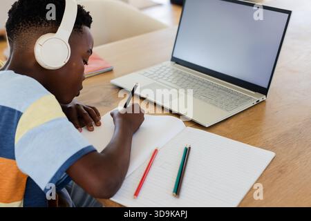 Afroamerikanischer Junge in gestreiftem Hemd, der im Notebook am Schreibtisch mit Laptop, Bleistift, Headset schreibt Stockfoto